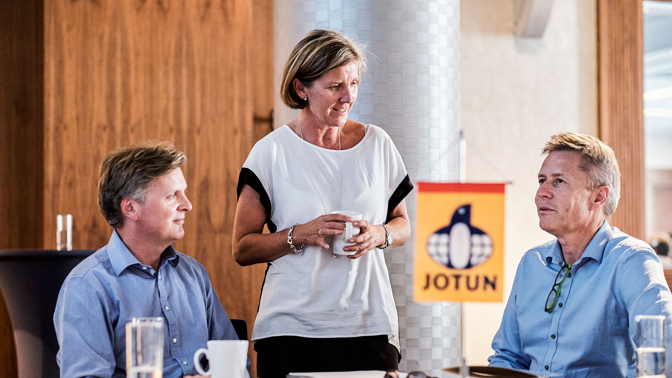 Two men with blue shirts sitting and a woman standing holding a coffee cup